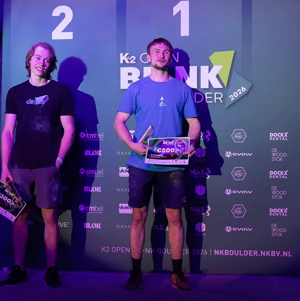 Twee mannelijke atleten staan op een podium: de ene op de eerste plaats, met een prijzengeldcheque en een trofee in zijn handen, en de andere op de tweede plaats. Op de achtergrond zijn de logo’s van het evenement en de sponsors van de K2 Open Bunk Boulde